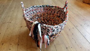 Av flerfärgade mattrasor har man slagit långa rep som sedan har sammanfogats med papperssnöre till en större rundformad, ca 50 cm hög kasse/korg med två handtag.