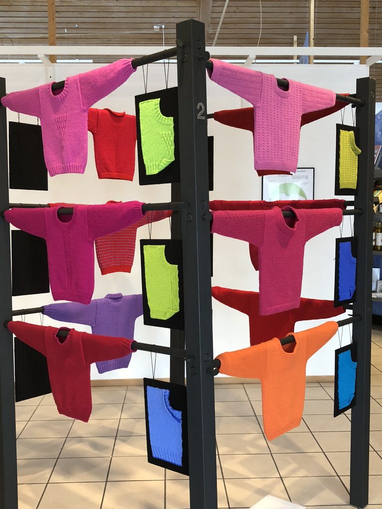 Utställning med stickade tröjor i starka rosa, röda. lila nyanser.