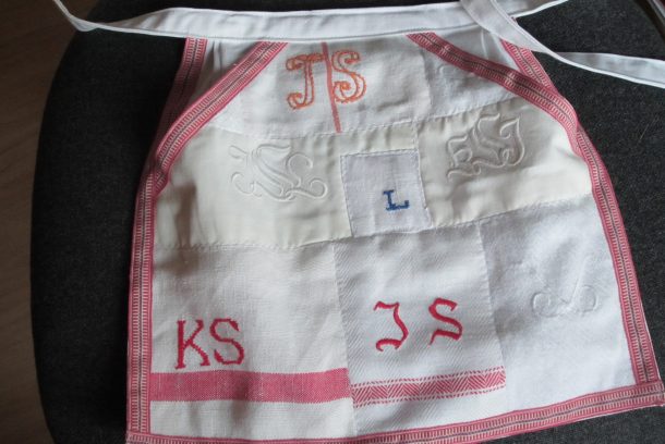 Litet förkläde i rött och vitt med olika initialer: JS, KS, L.