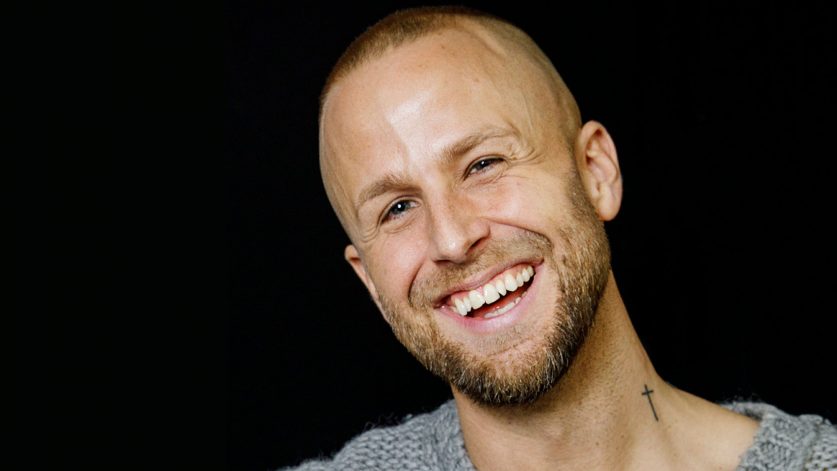 Are Norrhava ler i grå stickad tröja mot mörk bakgrund.